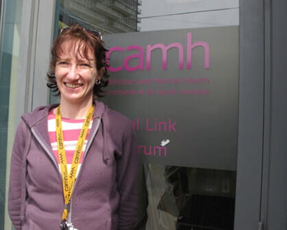A woman with ginger hair and bangs wearing a half zipped purple hoodie, smiling with her hands clasped behind her back.