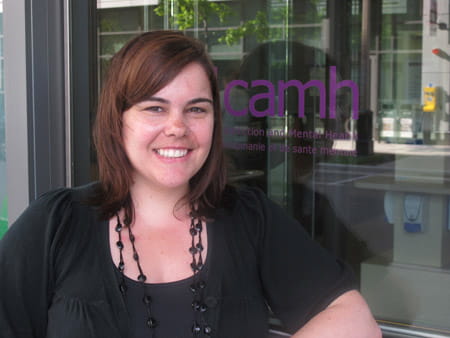 Cassandra wearing all black and a long black necklace posing beside the CAMH logo. 