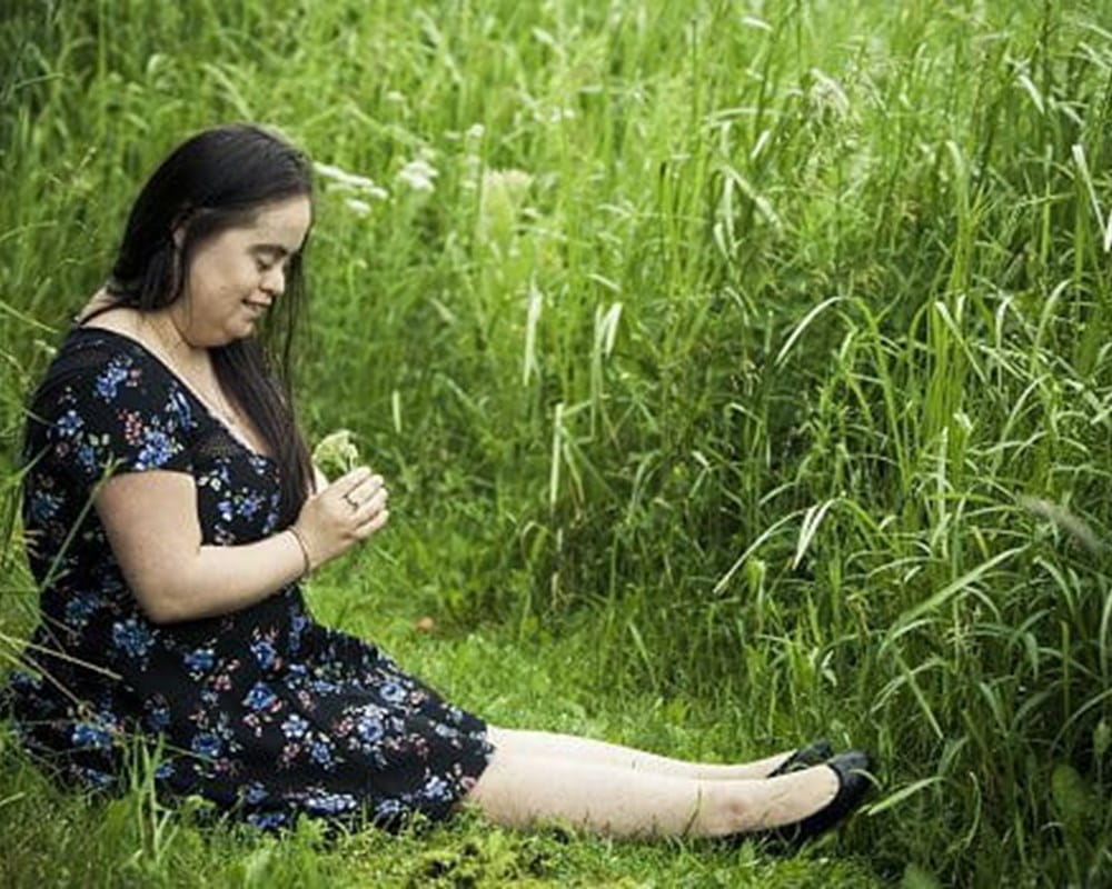 A woman with down syndrome wearing a black floral dress, sitting in a green field looking at a flower in her hand.