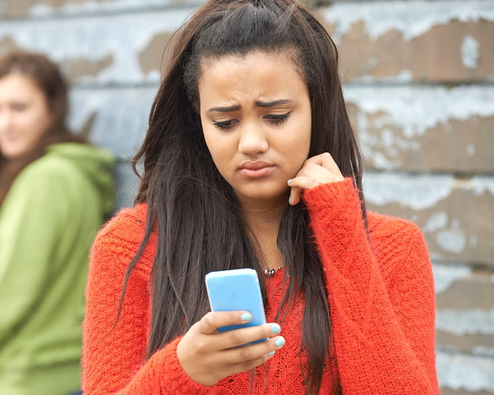Girls looks sadly at her phone with other girls gossiping in background