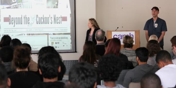 A woman presenting in front of a large diverse crowd with a projected image of a newspaper headline 