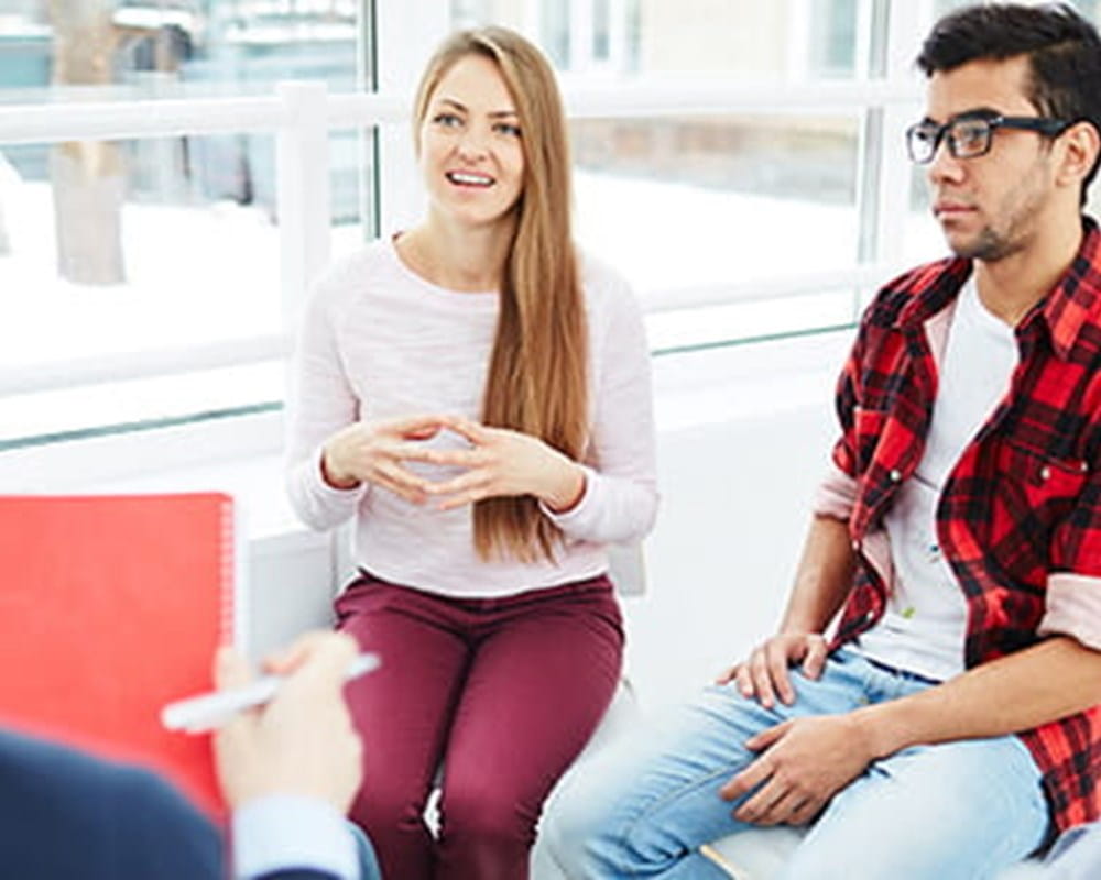 A woman with long hair and two men with facial hair sitting around a woman with classes and a red notebook.