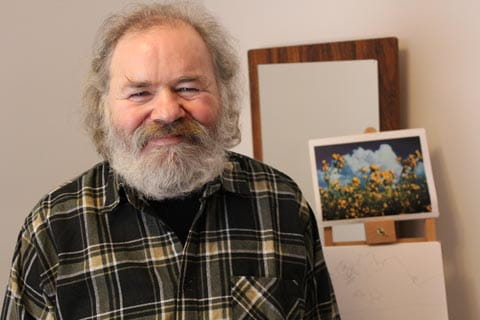An older man with grey hair and a grey beard in a plaid shirt smiling at the camera. 