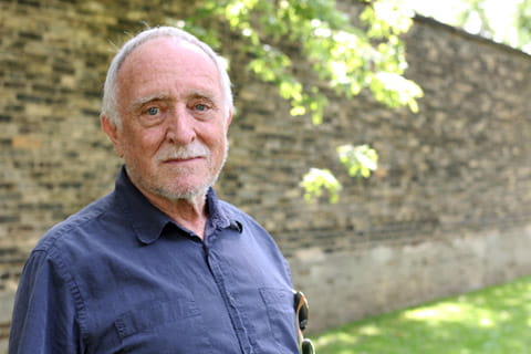 An older man with white hair wearing a blue button up standing in front of a brick wall, smiling for the camera. 