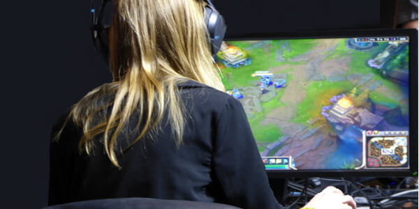 A person with medium length dirty blond hair wearing headphones sitting in front computer screen playing a video game. 