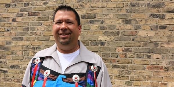 A man of Aboriginal descent smiling in front of a brick wall. 