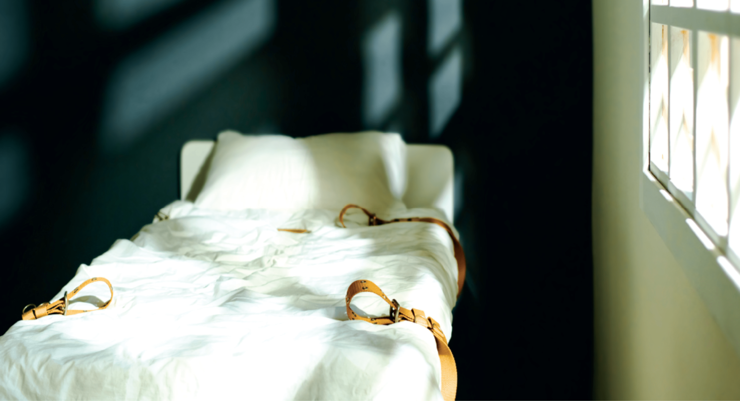 An empty white hospital bed with shackles next to a window. Shadows from the window frame cover the blank turquoise wall behind the bed. 